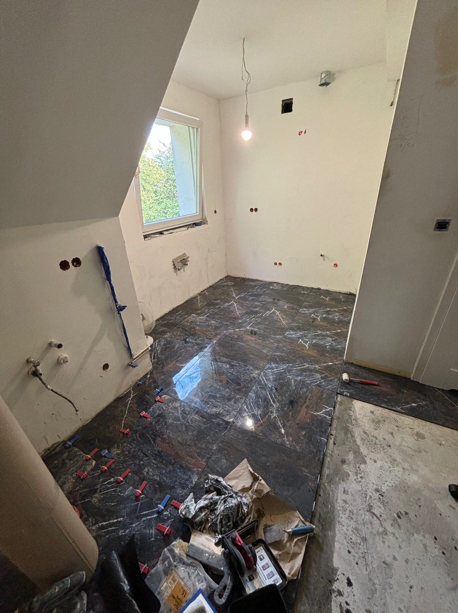 Układanie ciemnych płytek imitujących marmur w łazience na poddaszu, widoczne krzyżyki dystansowe, fragment okna, instalacja wodna i elektryczna w ścianach, narzędzia glazurnicze w dolnym rogu.