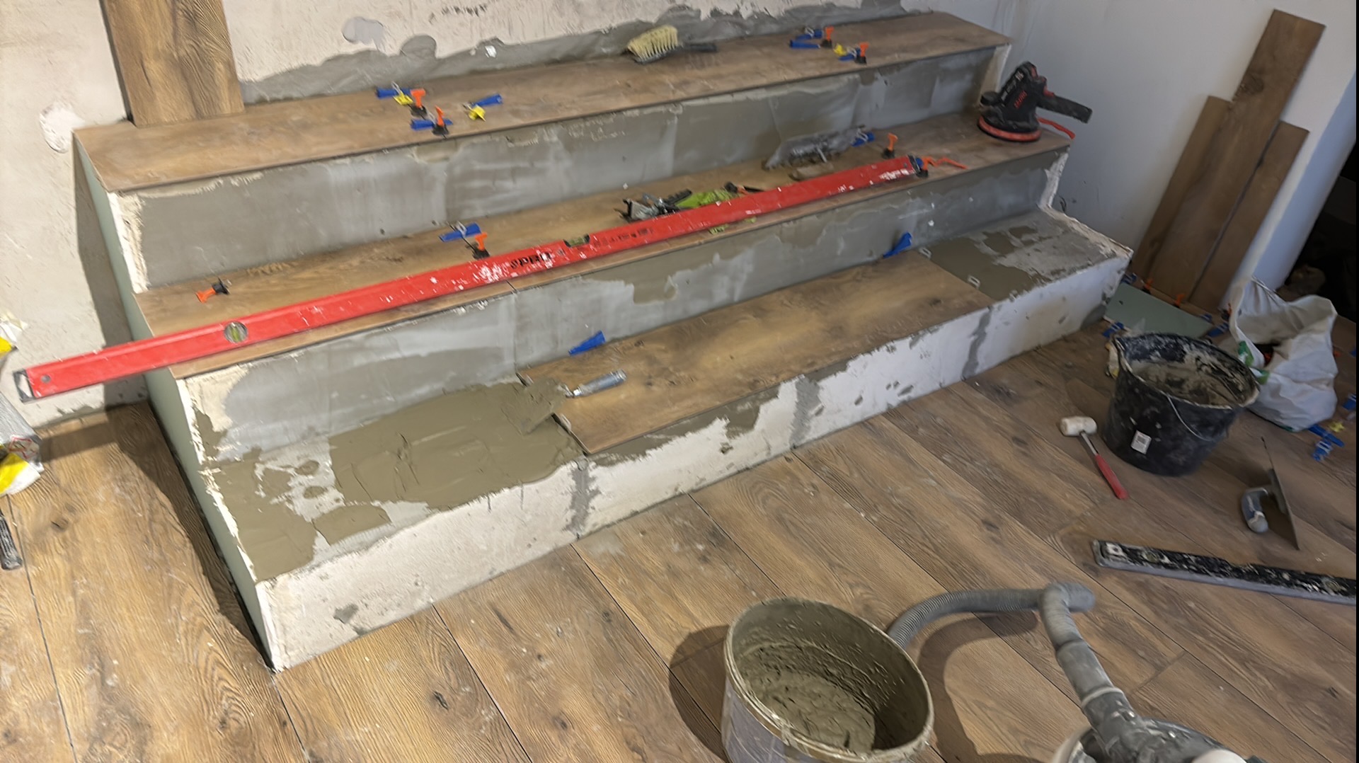 Układanie płytek imitujących drewno na schodach z użyciem poziomicy, kielni i systemu poziomowania, widoczne wiadro z zaprawą klejową i odkurzacz przemysłowy.