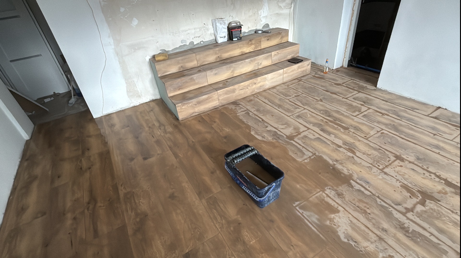 Układanie podłogi z płytek imitujących drewno, widoczna kuweta z wałkiem do fugowania i trójstopniowy podest obłożony tymi samymi płytkami, ściany w trakcie przygotowania.