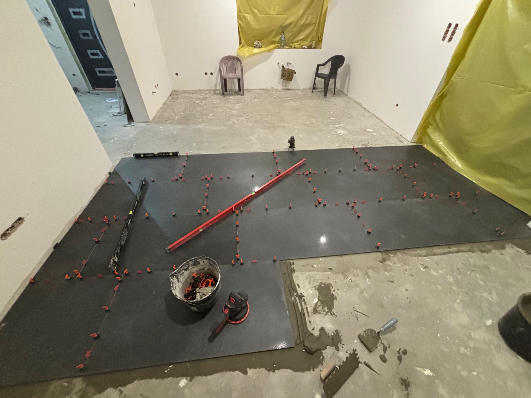 Układanie ciemnych płytek podłogowych z użyciem poziomicy, krzyżyków dystansowych i przyssawki do płytek, widoczny kubeł z zaprawą i szpachelka, w tle puste pomieszczenie z krzesłami i oknem...