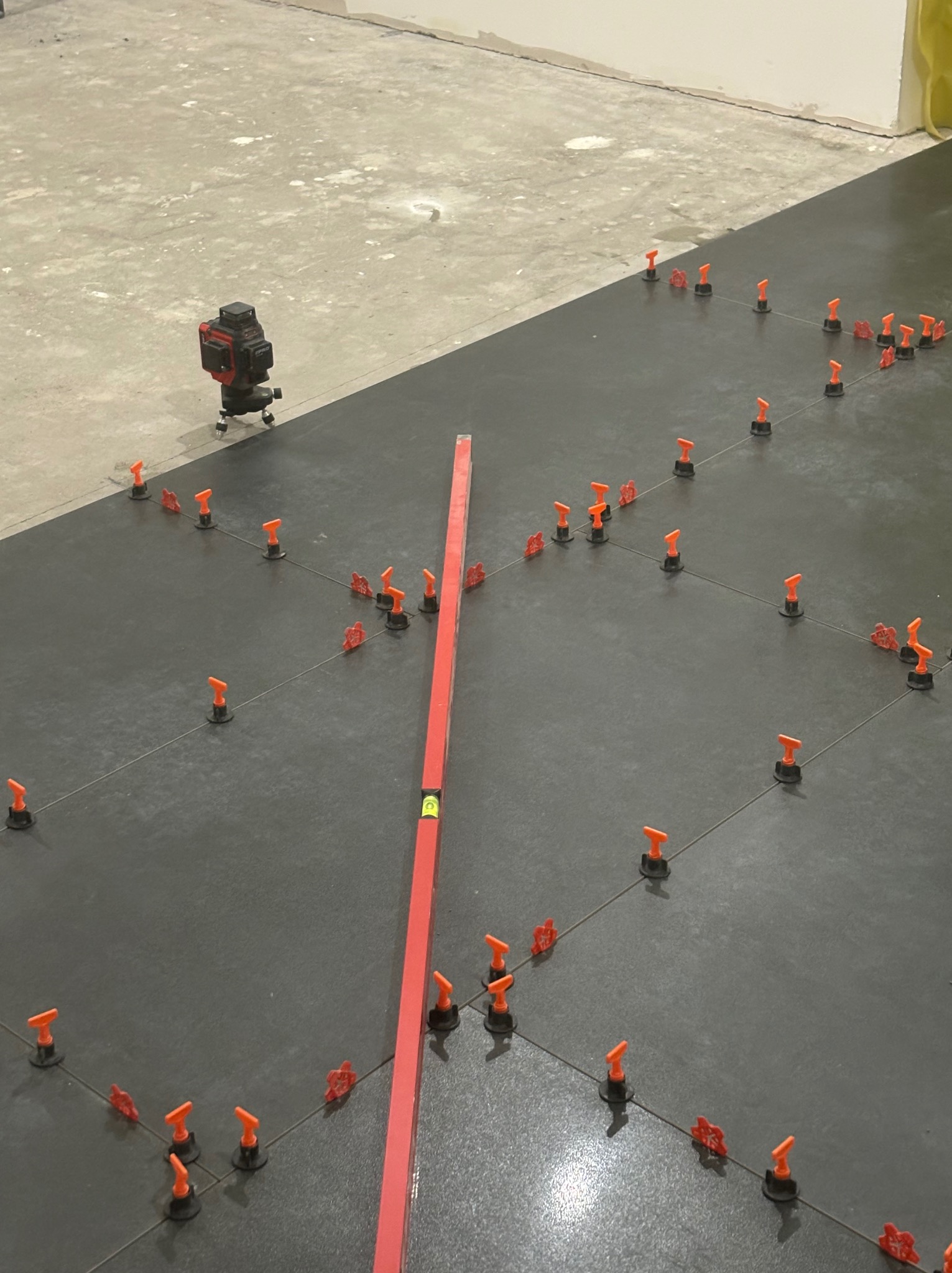 Proces układania ciemnych płytek podłogowych z użyciem poziomicy laserowej i pomarańczowych klipsów poziomujących.