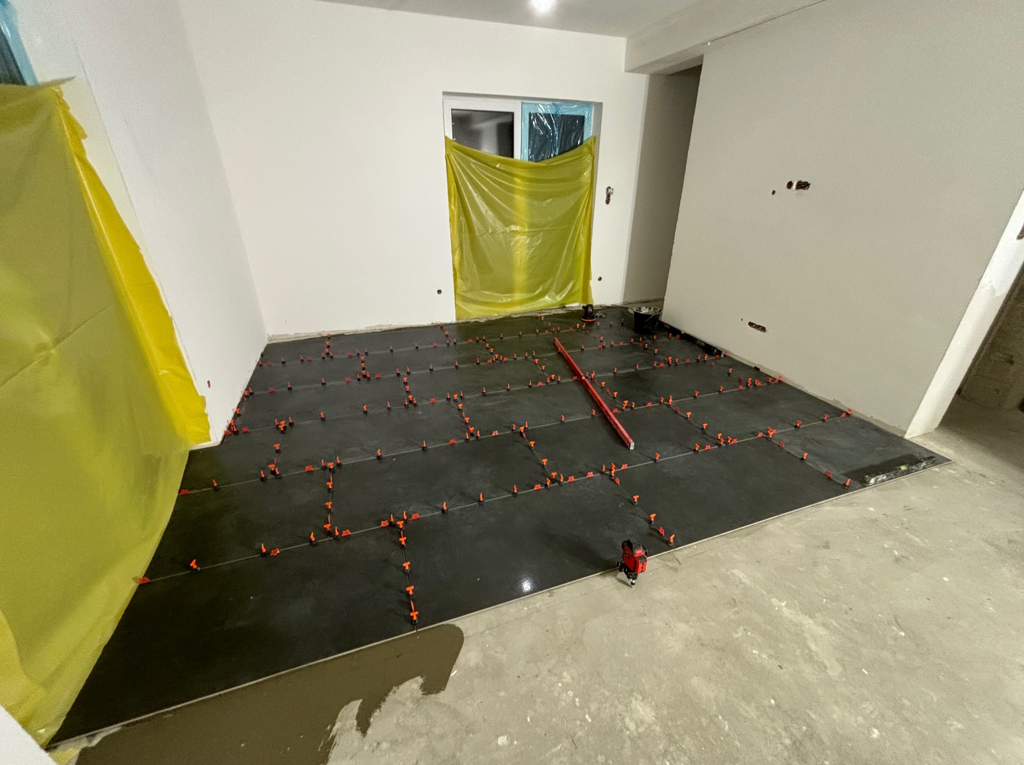Proces układania ciemnych płytek podłogowych z użyciem poziomicy laserowej i klipsów poziomujących, w pomieszczeniu zabezpieczonym folią malarską.