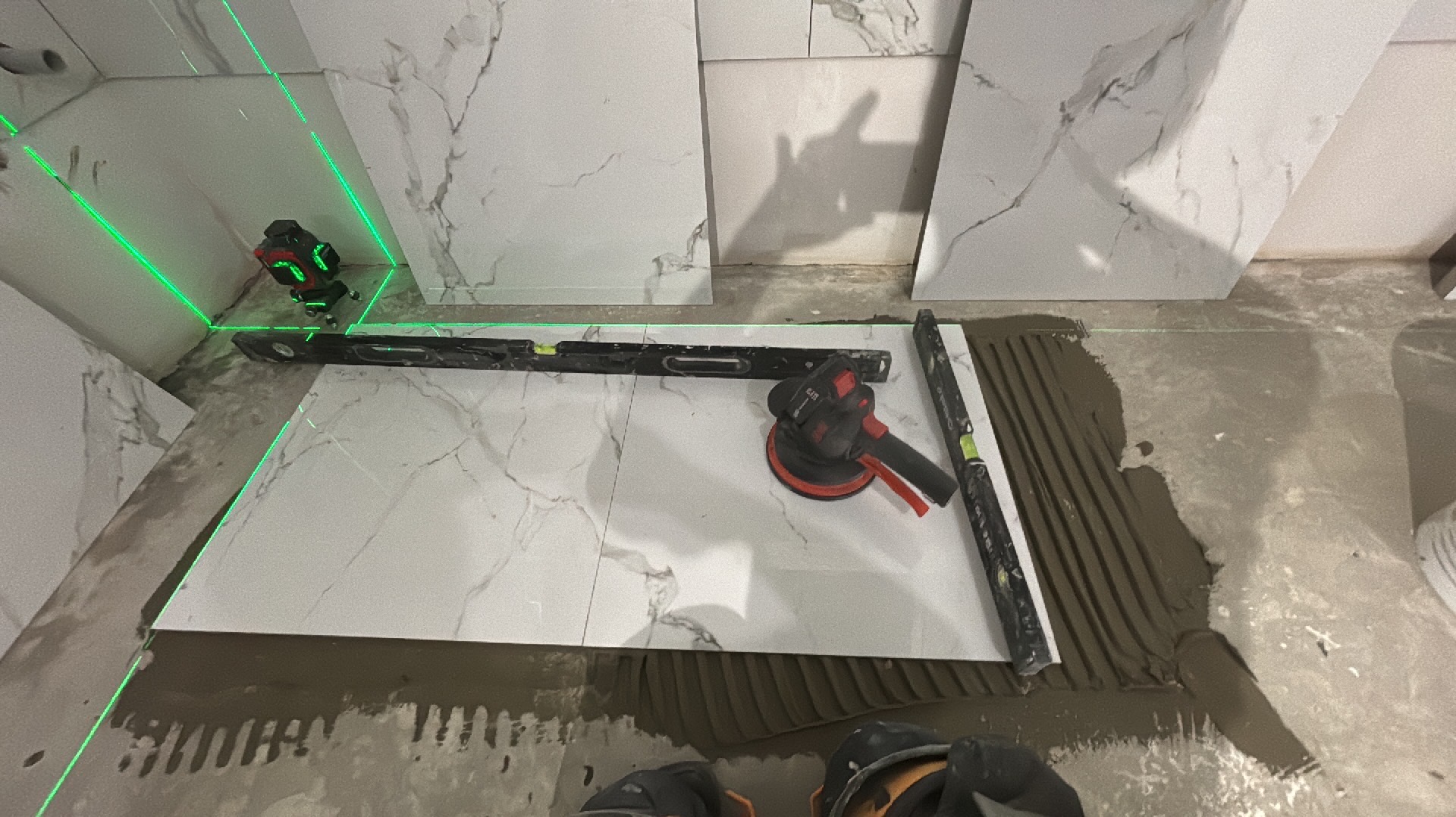 Układanie płytek imitujących marmur na podłodze, widoczny laser krzyżowy, poziomica, szlifierka do płytek i nałożony klej.