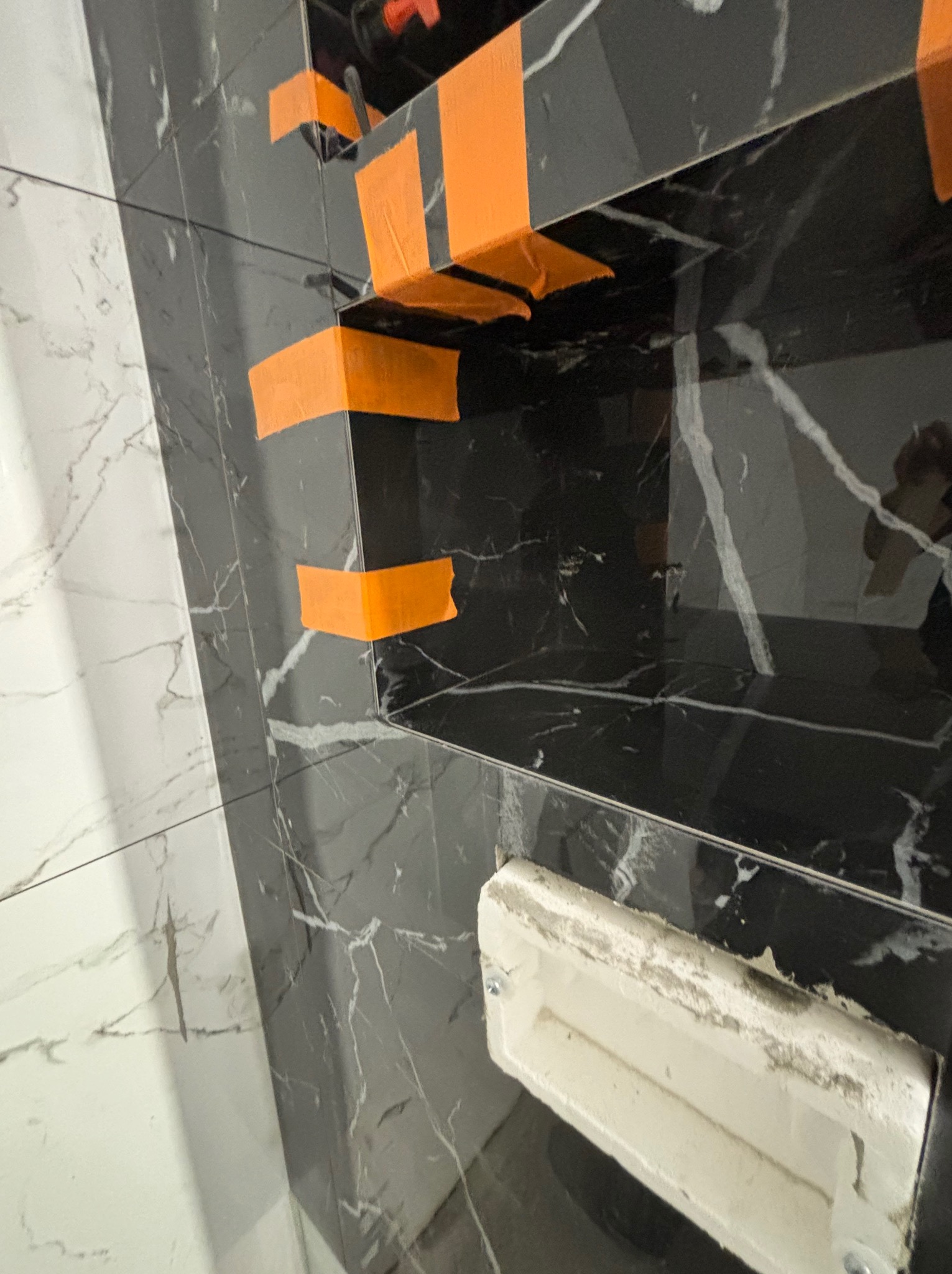 Fragment łazienki w trakcie wykańczania: czarne płytki imitujące marmur z pomarańczową taśmą maskującą, białe płytki obok i wnęka w ścianie z widocznym profilem montażowym.