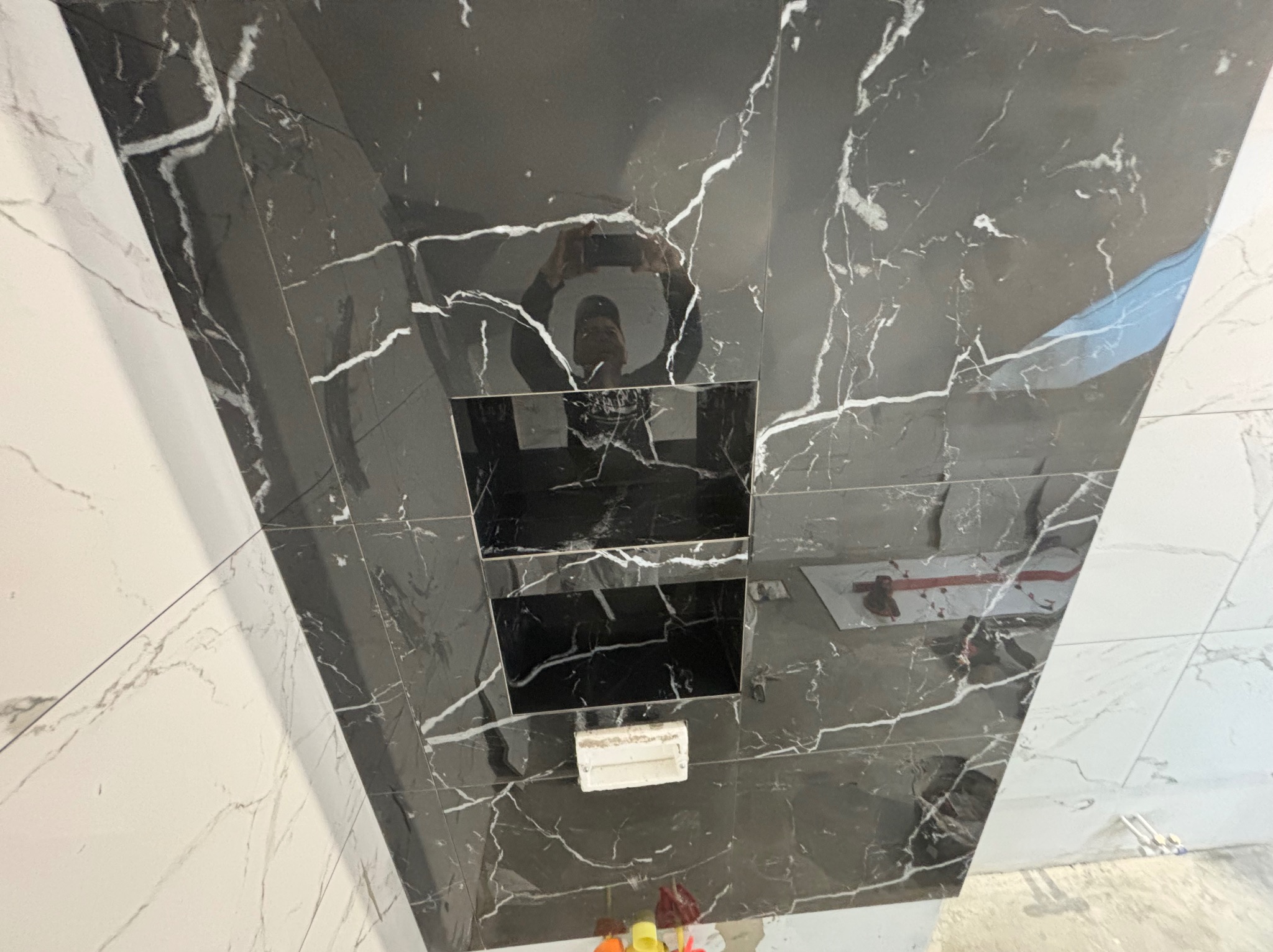 Ściana łazienki wyłożona czarnymi i białymi płytkami imitującymi marmur z wnękami na kosmetyki i odbiciem osoby robiącej zdjęcie.