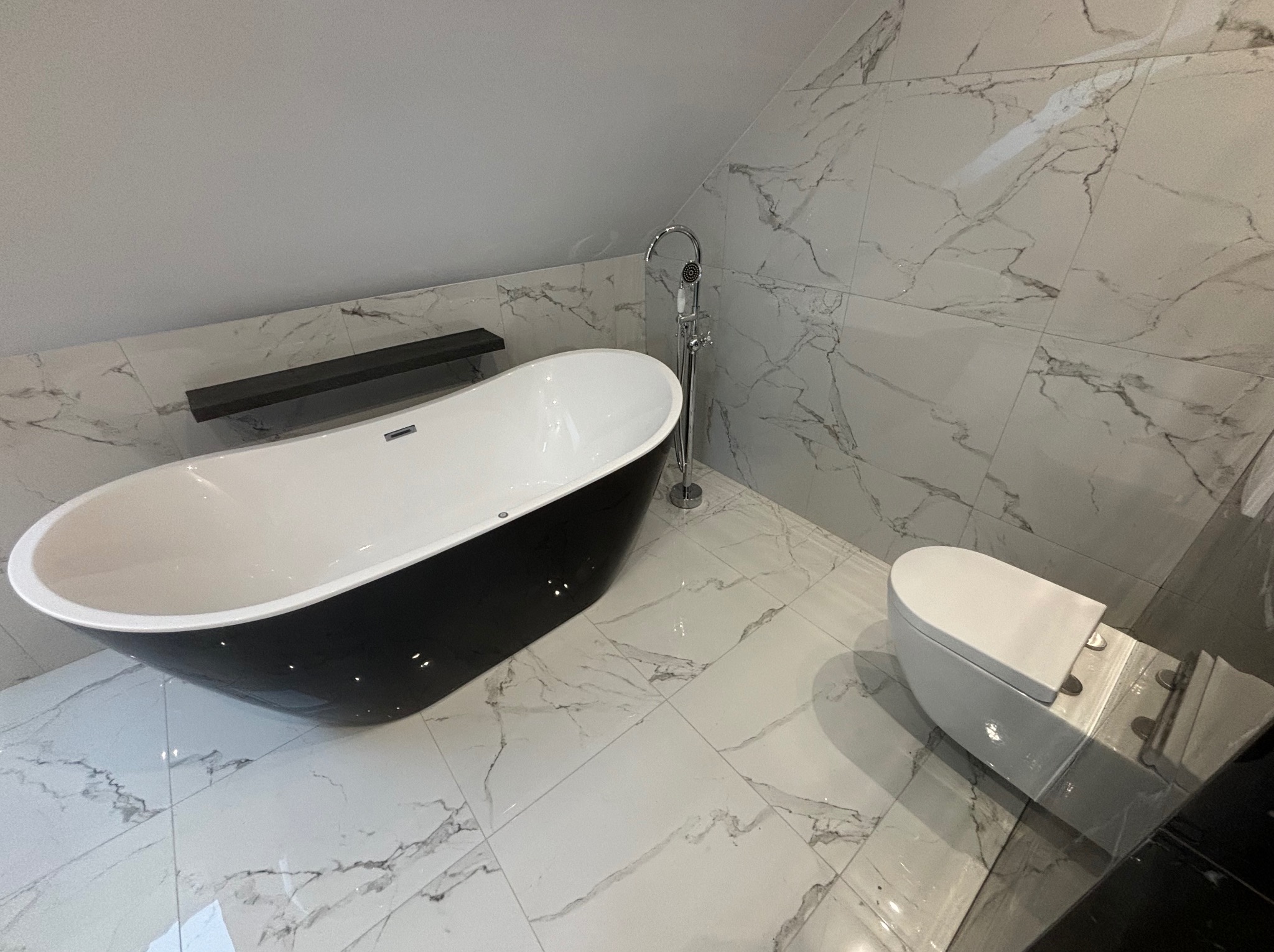 Nowoczesna łazienka na poddaszu z wolnostojącą czarno-białą wanną, toaletą typu Geberit i marmurowymi płytkami na ścianach i podłodze, oświetlona naturalnym światłem.