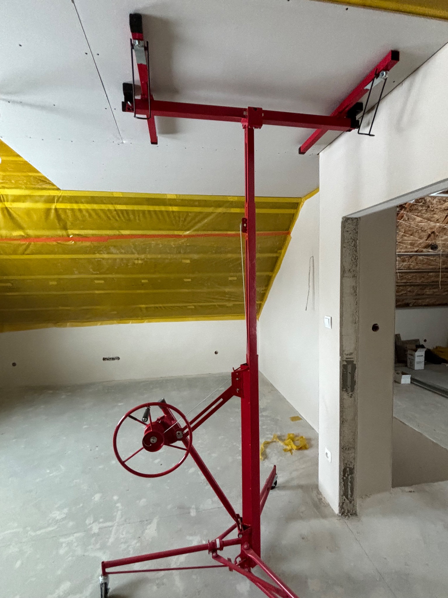 Czerwony podnośnik do płyt gipsowo-kartonowych podtrzymuje białą płytę sufitową, widoczne żółte zabezpieczenie folią, surowy stan wykończenia wnętrza