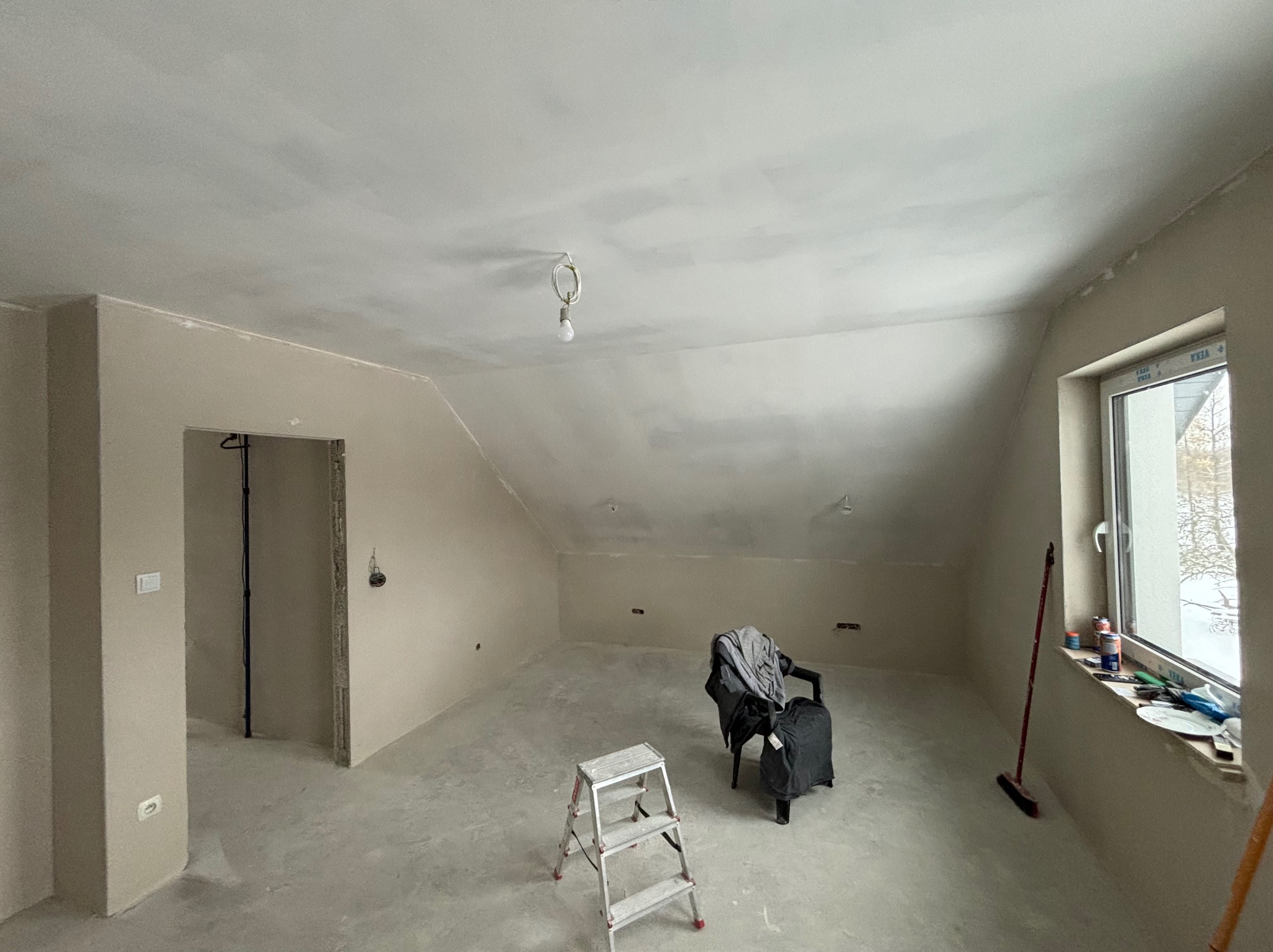 Poddasze po szpachlowaniu ścian i sufitu, widoczne ślady po szpachli, drabina, krzesło z ubraniami, okno z narzędziami i szczotką.