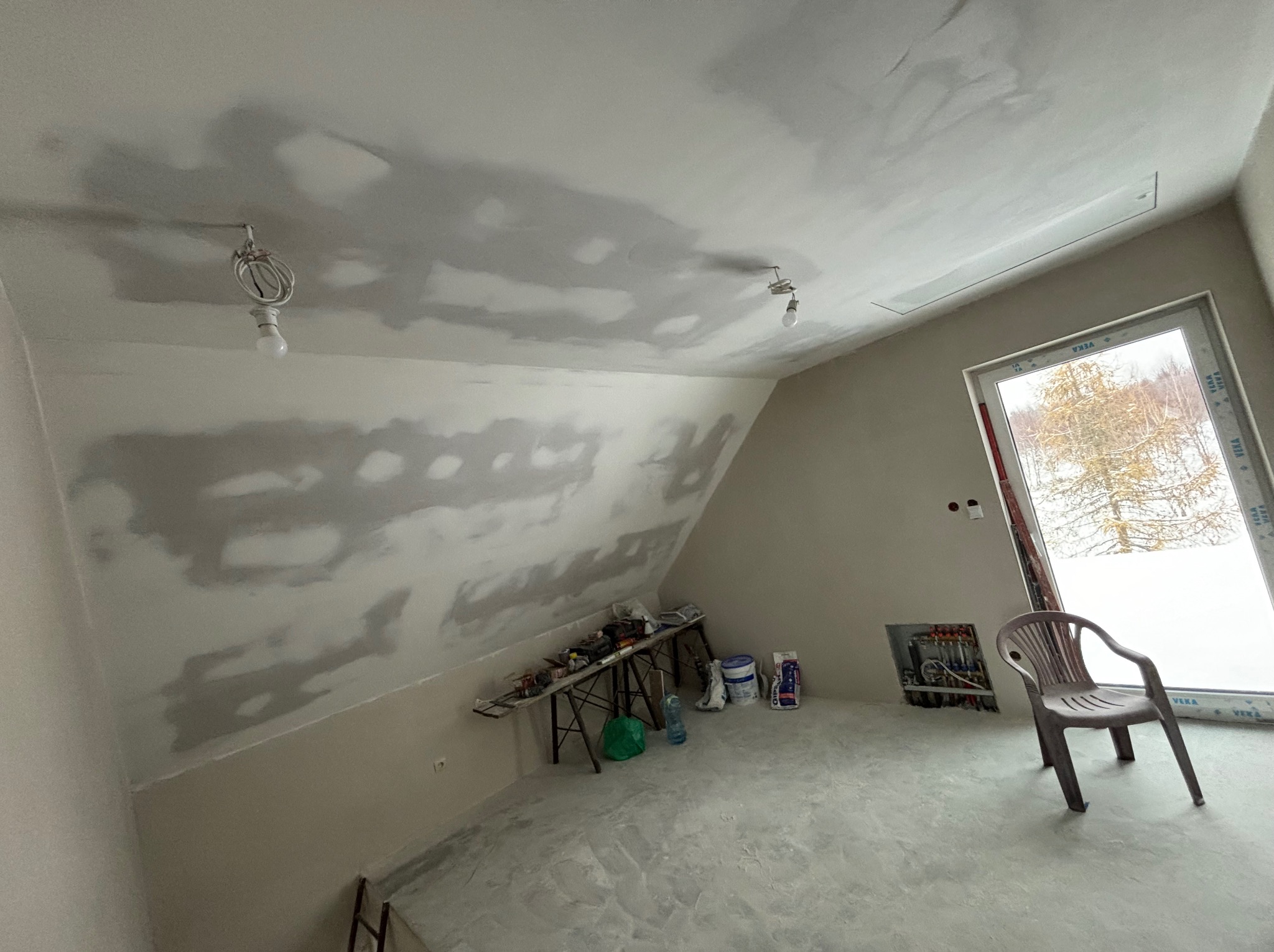 Poddasze w Koszalinie w trakcie szpachlowania, widoczne narzędzia na stole roboczym, odkryte przewody elektryczne oraz plastikowe krzesło przy oknie.
