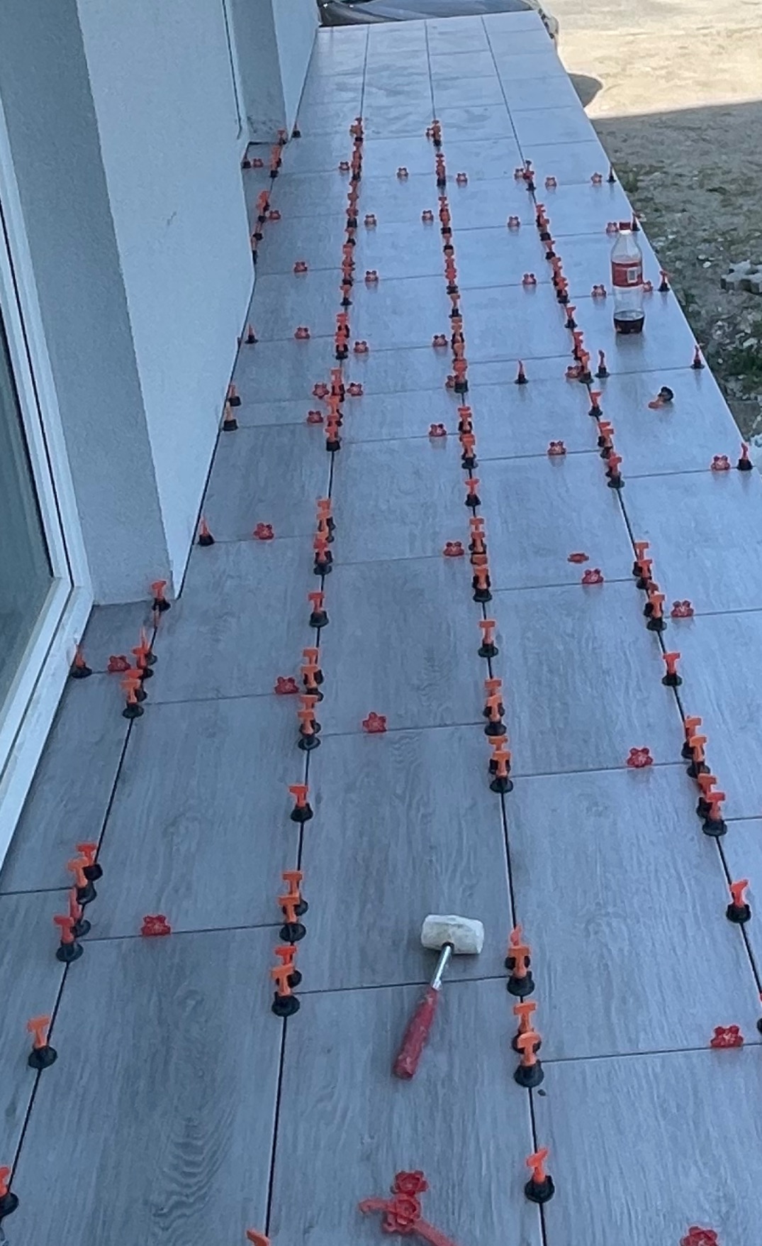 Układanie szarych płytek na zewnątrz budynku, widoczne krzyżyki dystansowe, kliny poziomujące, sznurek traserski, gumowy młotek i butelka napoju.