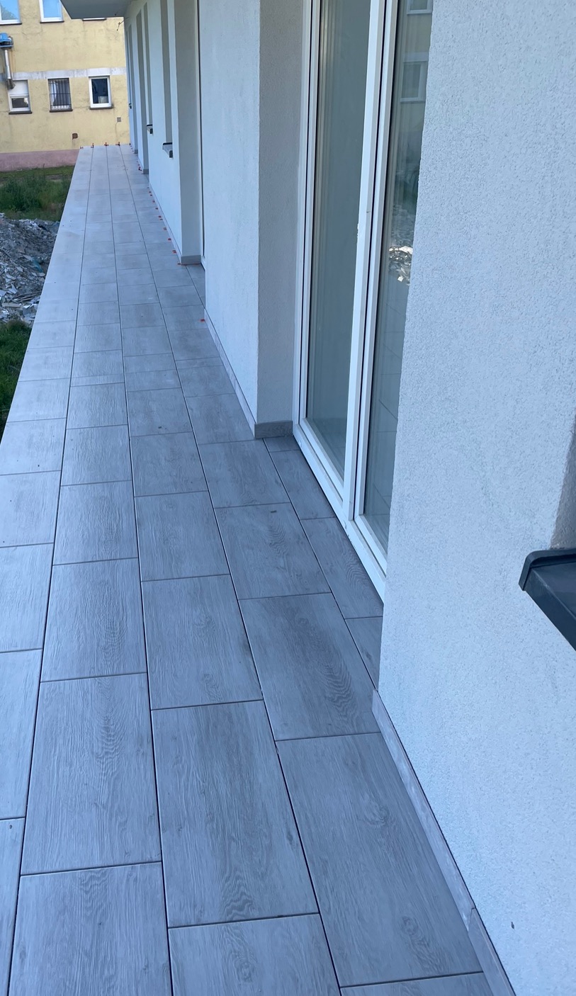 Wyłożony jasnoszarą płytką imitującą drewno balkon w nowym budownictwie, widoczne białe ramy okienne i fragment elewacji.