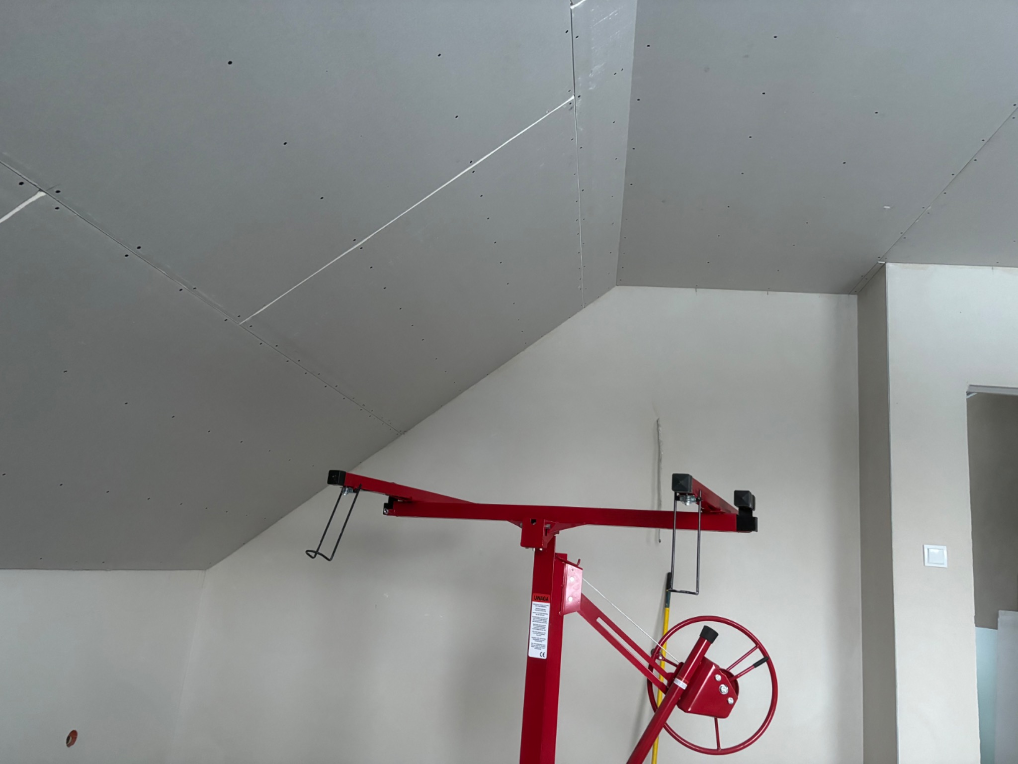 Montaż płyt kartonowo-gipsowych na skośnym suficie poddasza z użyciem czerwonego podnośnika, widoczne łączenia płyt i otwory po wkrętach.