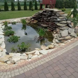 Oczko wodne z kaskadą z kamieni łupkowych, otoczone roślinnością wodną i dekoracyjnymi kamieniami, z drewnianym domkiem na drzewie w tle, widoczne z poziomu brukowanego chodnika.