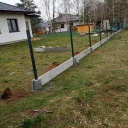Montaż betonowej podmurówki i słupków metalowych pod ogrodzenie panelowe na trawiastym terenie, widoczny dom w tle.