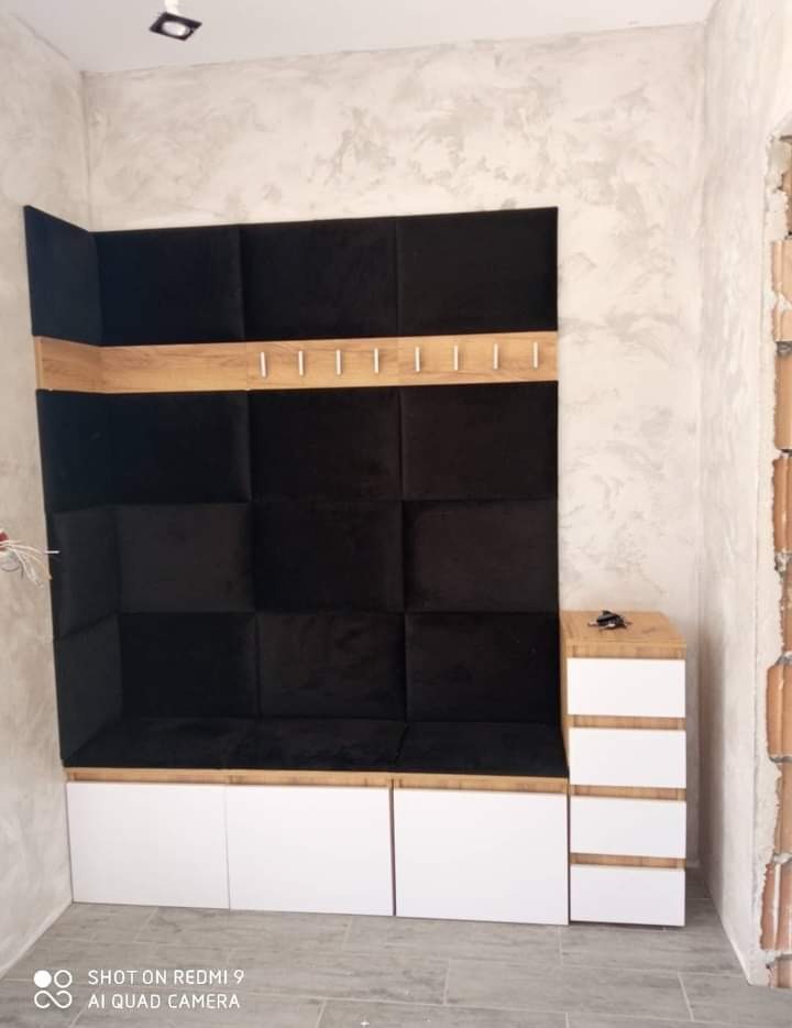 Wnęka z siedziskiem wykończona czarnymi, tapicerowanymi panelami, drewnianą półką z wieszakami i białymi szafkami, surowy tynk na ścianie