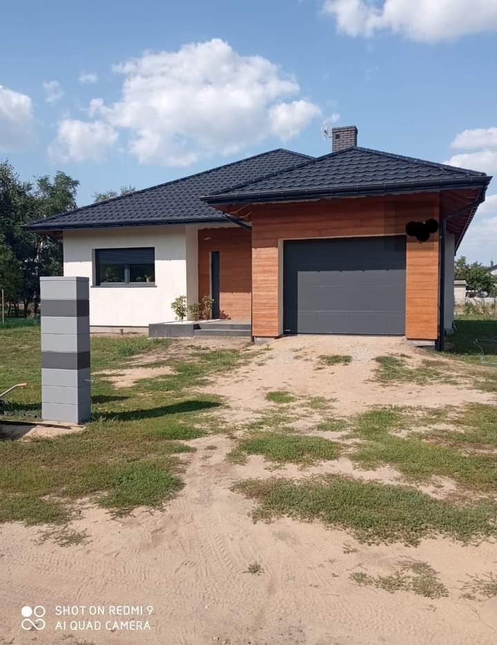 Nowoczesny dom jednorodzinny z grafitową bramą garażową i drewnianą elewacją, widoczny z zewnątrz na tle błękitnego nieba z chmurami.