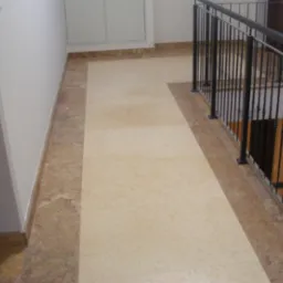 Korytarz wykończony jasnym kamieniem z ciemniejszym obramowaniem, widoczne schody z czarną balustradą i drzwi.