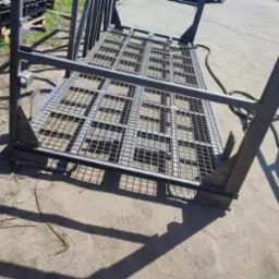 Metalowa konstrukcja kratowa, prawdopodobnie wózek lub rama, po malowaniu proszkowym na szaro, widoczne spawy, na betonowym podłożu z elementami roślinności w tle.