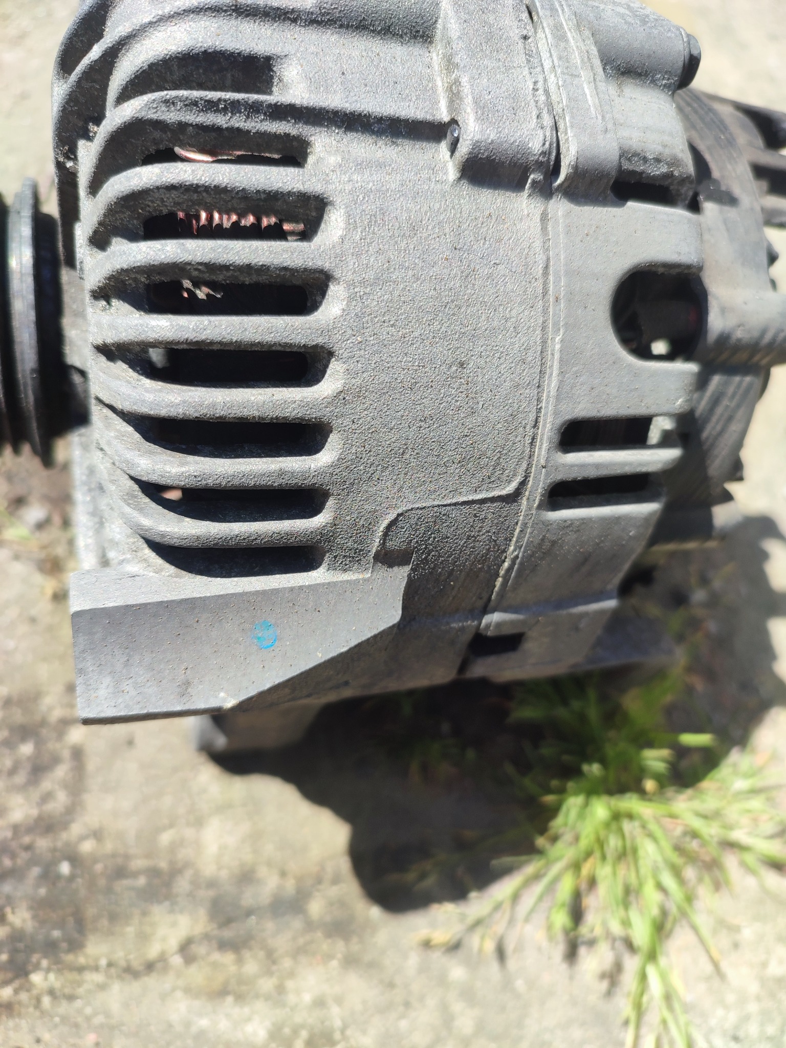 Szary alternator samochodowy z widocznymi żebrami chłodzącymi i śladami użytkowania, leżący na betonowej powierzchni z kępką trawy w tle, z niebieską kropką farby na obudowie.