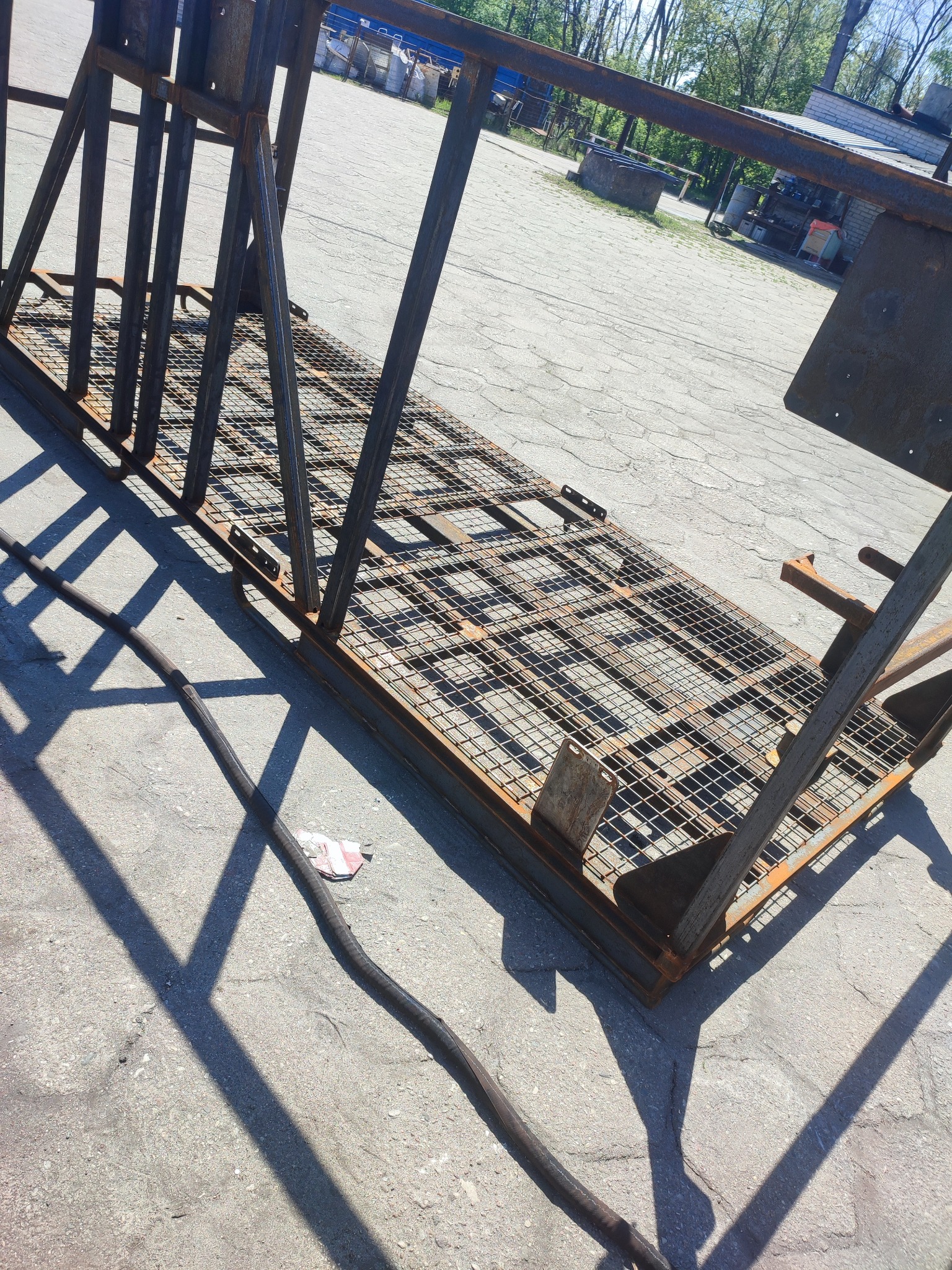 Metalowa konstrukcja kratowa z widocznymi śladami rdzy, prawdopodobnie przed procesem czyszczenia, z przewodem na betonowym podłożu i cieniem rzucanym przez konstrukcję.