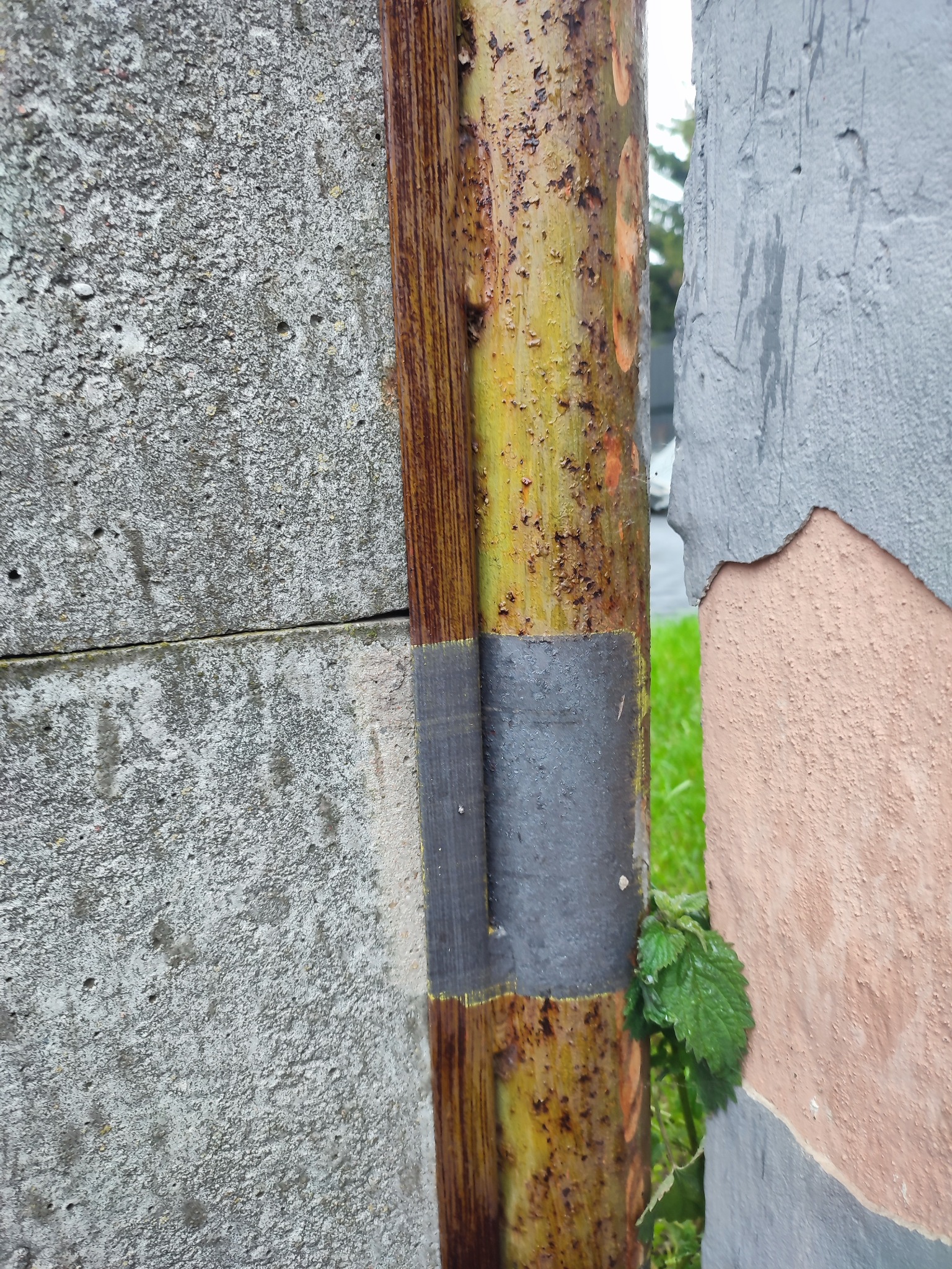 Zardzewiała metalowa rura z pozostałościami starej farby, owinięta taśmą, przylegająca do betonowego muru i ściany z odpadającym tynkiem w odcieniach szarości i brzoskwini, z widocznym fragmentem...