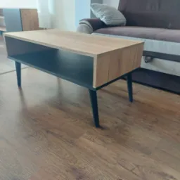 Stolik kawowy z drewnianym blatem i czarnymi nogami, z otwartą przestrzenią pod blatem, na tle brązowej kanapy i drewnianej podłogi.