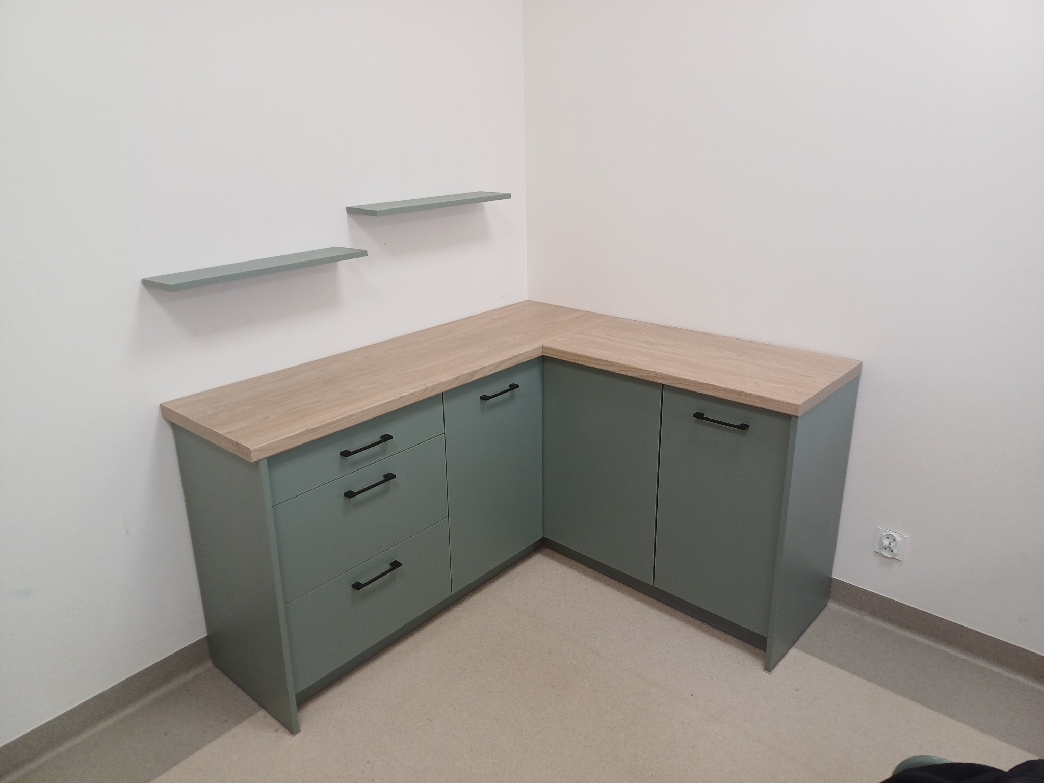 Narożna zabudowa kuchenna w odcieniach zieleni z drewnianym blatem, minimalistyczne czarne uchwyty, dwie półki wiszące na ścianie powyżej, widok z dołu.