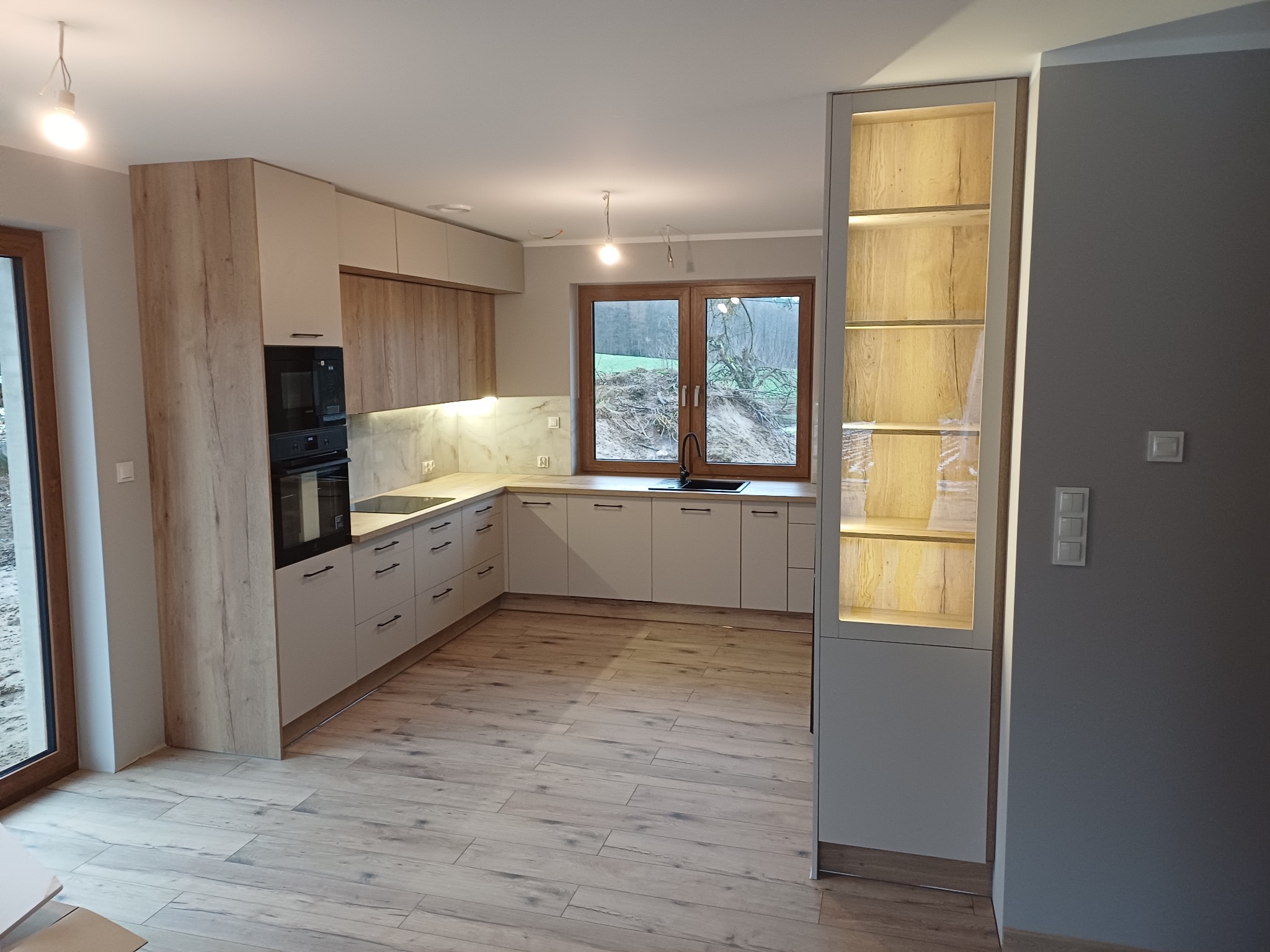 Jasna kuchnia w stylu skandynawskim z drewnianymi akcentami: fronty szafek z wyraźnym rysunkiem drewna, marmurowy backsplash, drewniana podłoga, okno z widokiem na zieleń, oświetlona witryna...