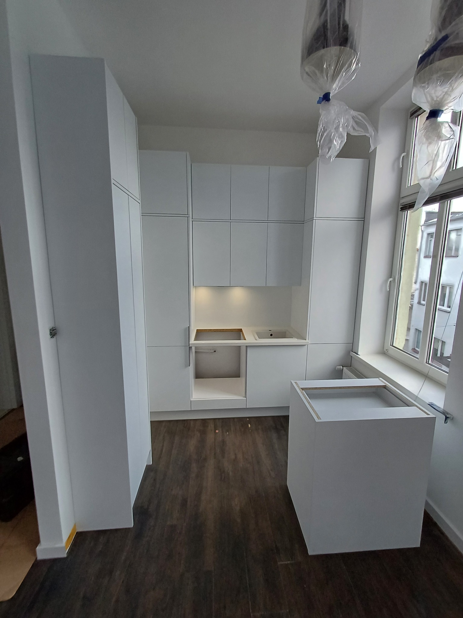 Montaż białej kuchni: widok na zabudowę kuchenną z szafkami wiszącymi i stojącymi, fragment wyspy kuchennej bez blatu, okno z widokiem na budynek, zabezpieczone lampy wiszące folią ochronną.