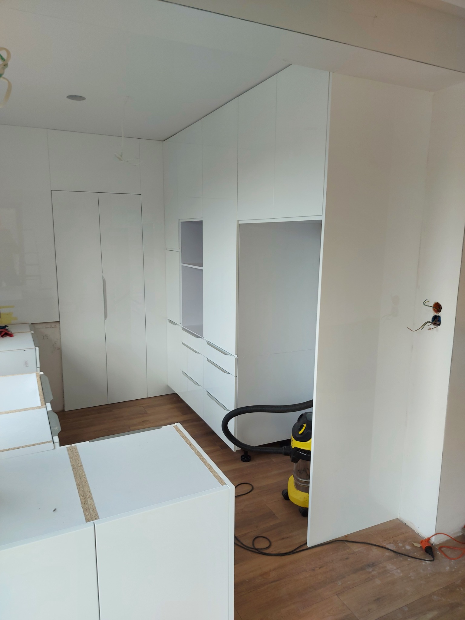 Montaż białej szafy wnękowej z szufladami i półkami, widoczne elementy do montażu, odkurzacz na podłodze, surowa ściana z wystającymi kablami elektrycznymi.