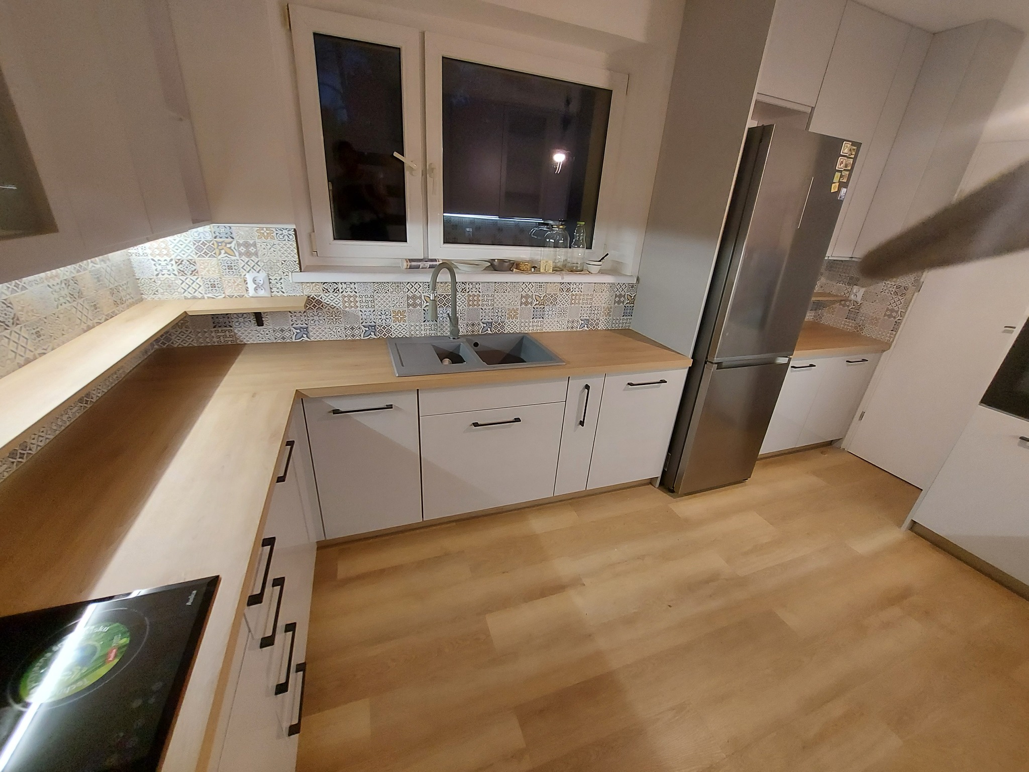 Jasna kuchnia z drewnianym blatem, szarym zlewozmywakiem i wzorzystą mozaiką nad blatem, widziana z perspektywy podłogi.