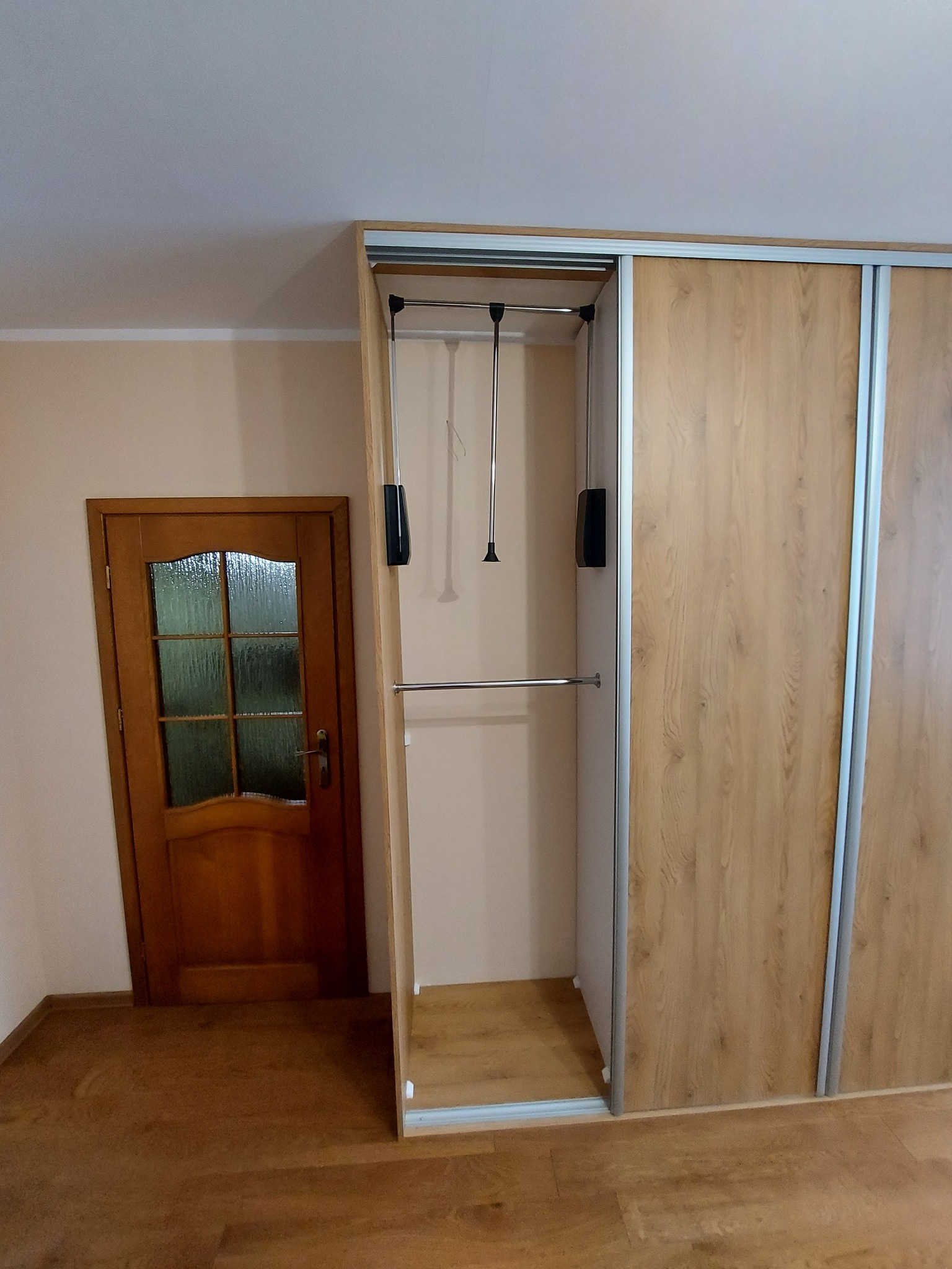 Wnętrze szafy przesuwnej z drewnopodobnymi frontami, widoczny drążek i pantograf, obok drewniane drzwi z szybą. Podłoga wykończona panelami w odcieniach brązu.