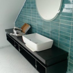 Jarosław Radziszewski - Nowoczesna łazienka w trakcie wykańczania: turkusowe płytki na ścianie, owalne lustro z podświetleniem, biała umywalka na czarnej szafce, biała podłoga w jodełkę.