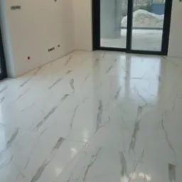 Położona, błyszcząca, biała podłoga z marmurowym wzorem w nowym, jasnym pokoju z dużym oknem przesuwnym i nowoczesną lampą sufitową.