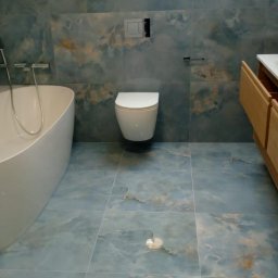 Jarosław Radziszewski - Wykończona łazienka z niebieskimi płytkami imitującymi marmur, wisząca toaleta, wolnostojąca wanna i szafka z drewnianymi frontami. Widoczne okno dachowe.