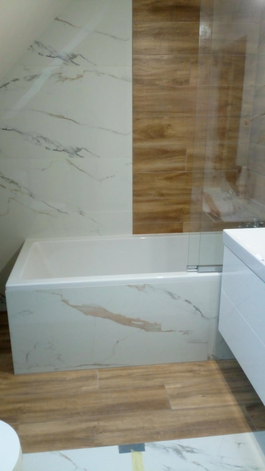 Wykończona łazienka z wanną obudowaną płytkami imitującymi marmur, drewnopodobna podłoga i ściana z płytek w dwóch odcieniach. Kabina prysznicowa ze szkła.