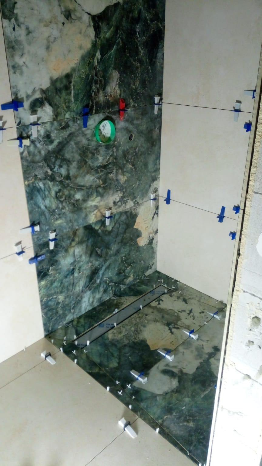 Układanie płytek w łazience z użyciem systemu poziomowania. Widoczne płytki imitujące marmur w odcieniach zieleni i beżu oraz elementy systemu poziomowania płytek.