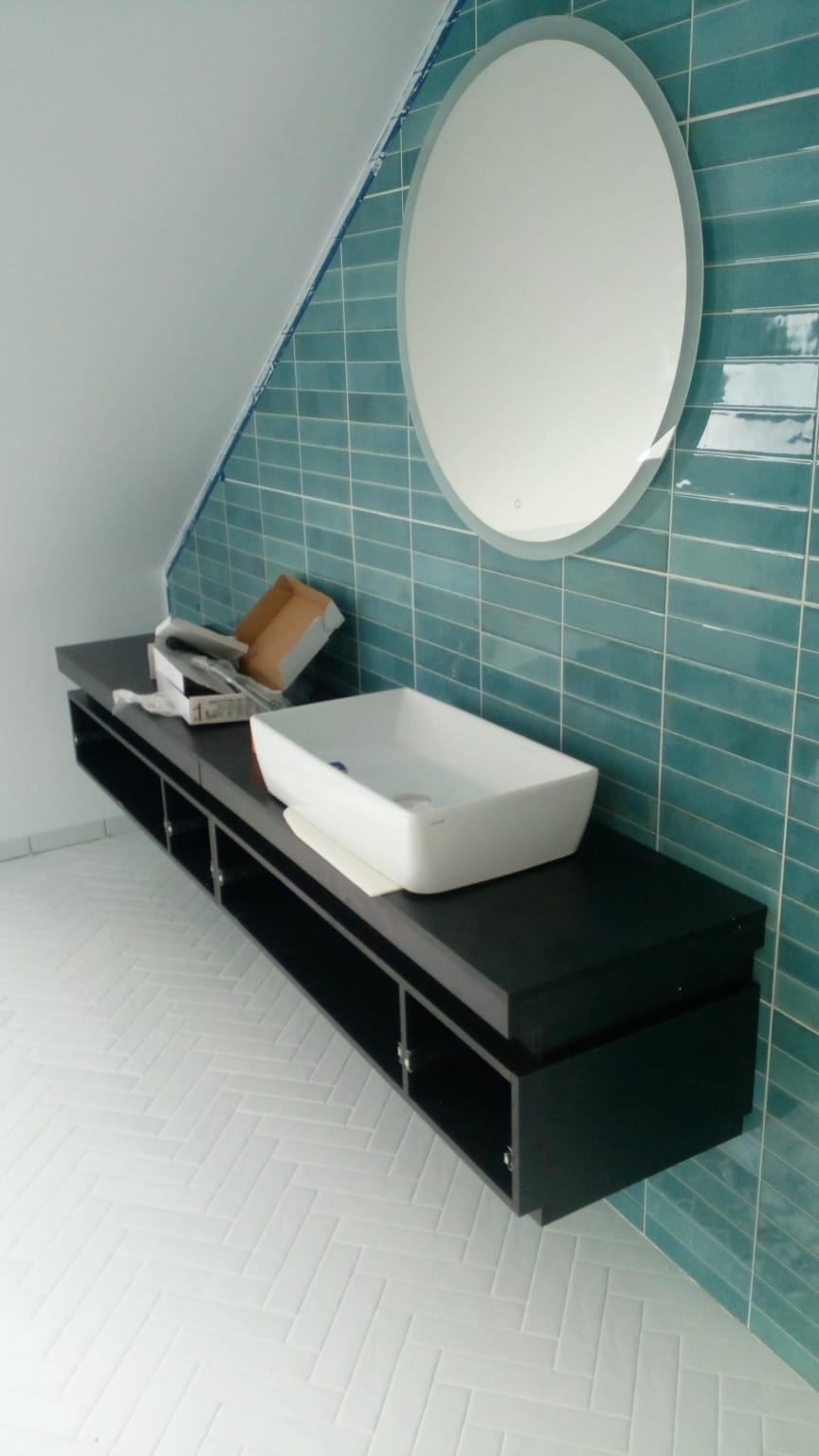 Nowoczesna łazienka w trakcie wykańczania: turkusowe płytki na ścianie, owalne lustro z podświetleniem, biała umywalka na czarnej szafce, biała podłoga w jodełkę.