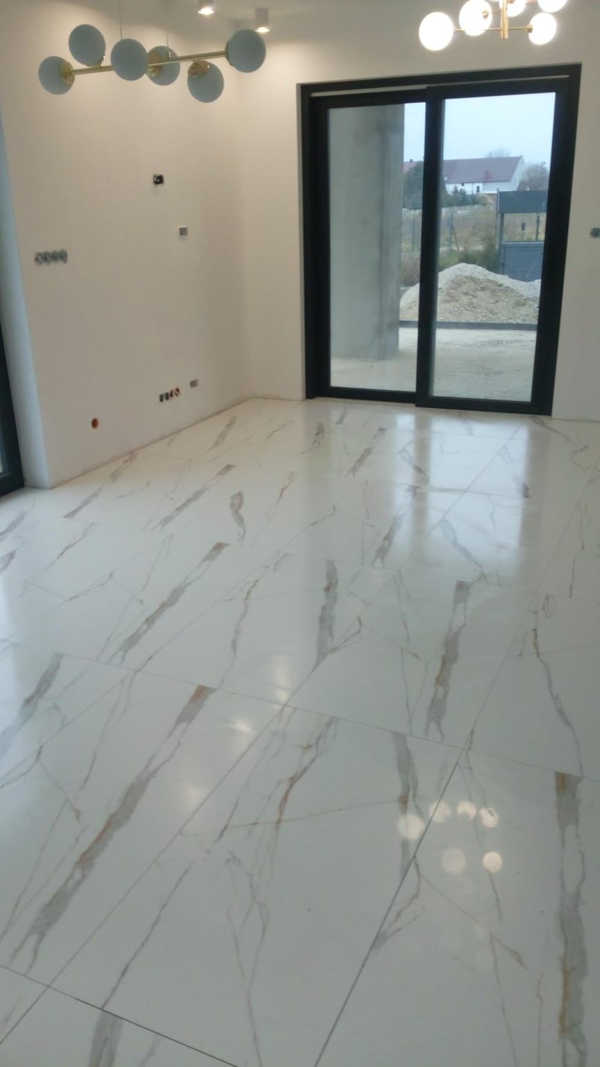 Położona, błyszcząca, biała podłoga z marmurowym wzorem w nowym, jasnym pokoju z dużym oknem przesuwnym i nowoczesną lampą sufitową.