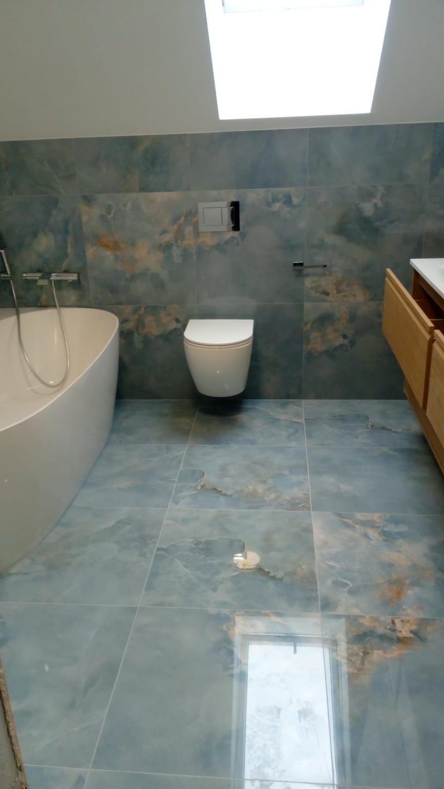 Wykończona łazienka z niebieskimi płytkami imitującymi marmur, wisząca toaleta, wolnostojąca wanna i szafka z drewnianymi frontami. Widoczne okno dachowe.