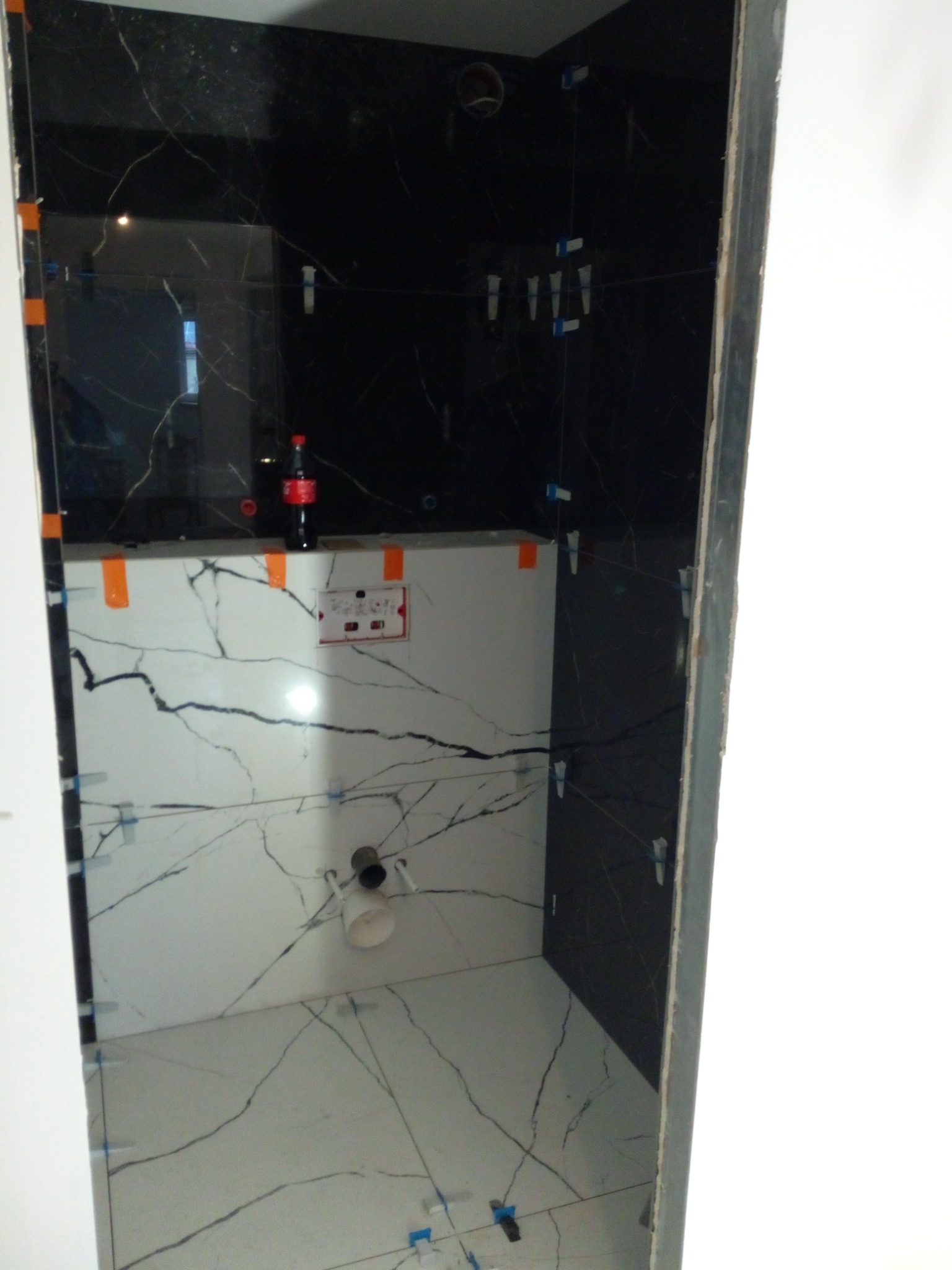 Łazienka w trakcie kładzenia płytek: czarne płytki imitujące marmur na ścianach, białe płytki z czarnymi żyłami na podłodze i obudowie stelaża podtynkowego, plastikowe krzyżyki dystansowe pomiędzy...