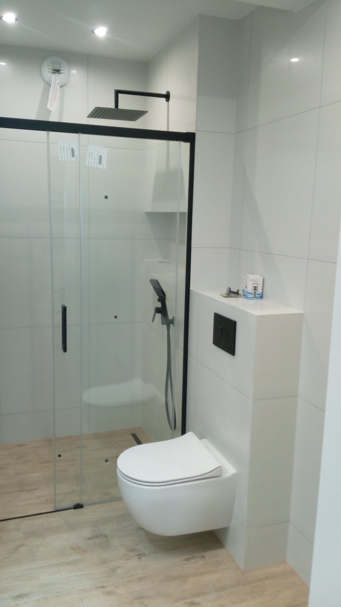 Nowoczesna łazienka z kabiną prysznicową typu walk-in z czarnymi ramami, wiszącą toaletą i drewnopodobnymi płytkami podłogowymi, minimalistyczny design.