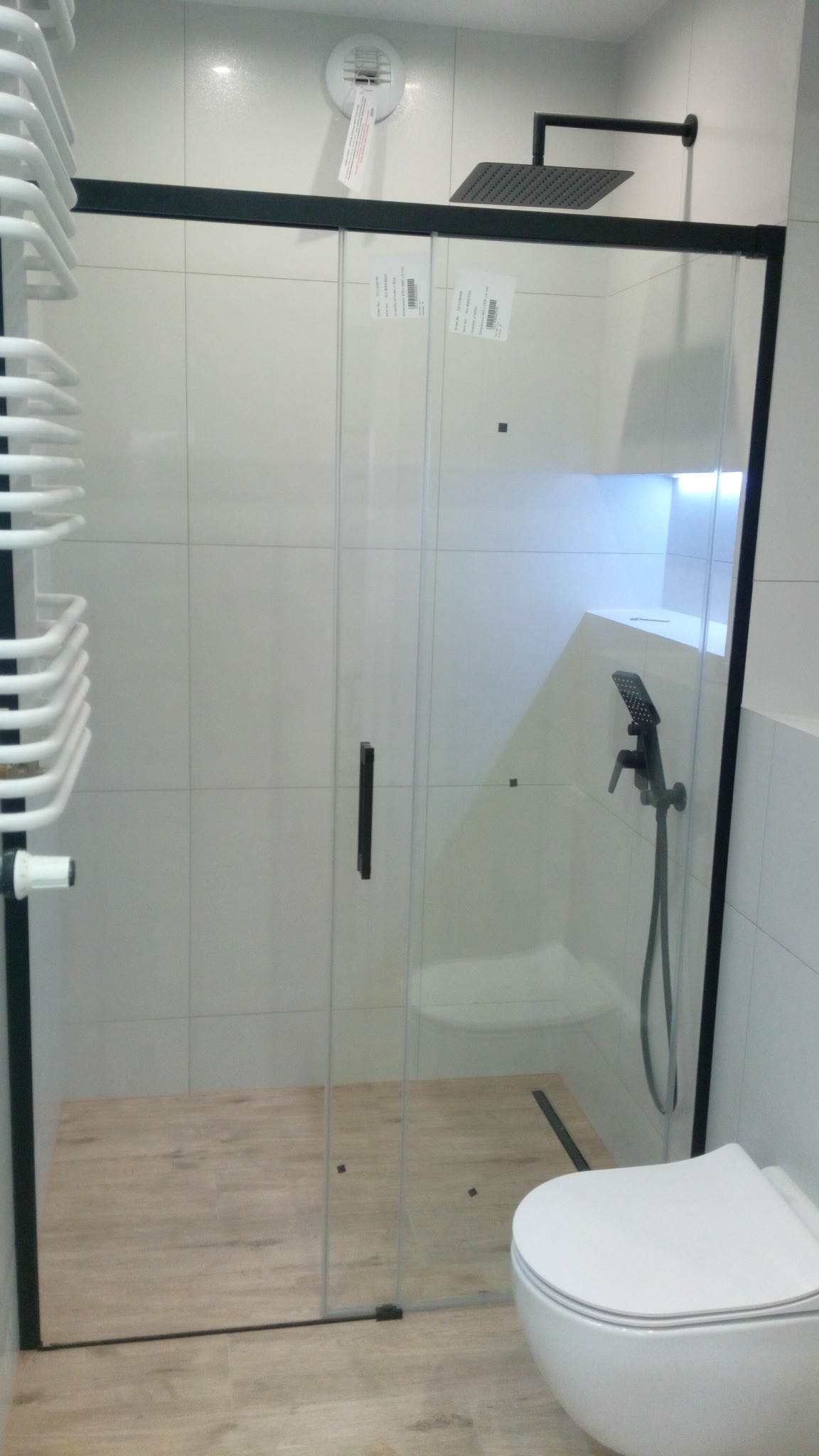 Nowoczesna kabina prysznicowa z czarnymi ramami i deszczownicą, widoczna toaleta wisząca i grzejnik drabinkowy, płytki imitujące drewno na podłodze.