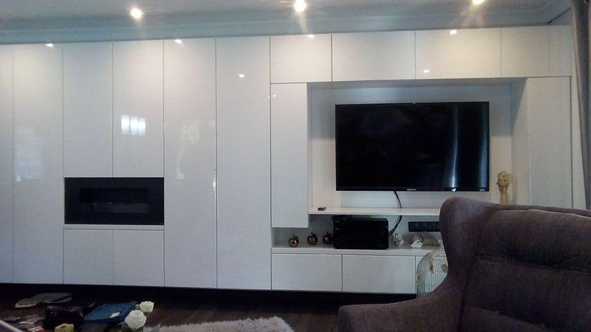 Biała, lakierowana szafa na wymiar z wnęką na telewizor i kominkiem elektrycznym, widok z perspektywy salonu, z fragmentem fotela w kadrze.