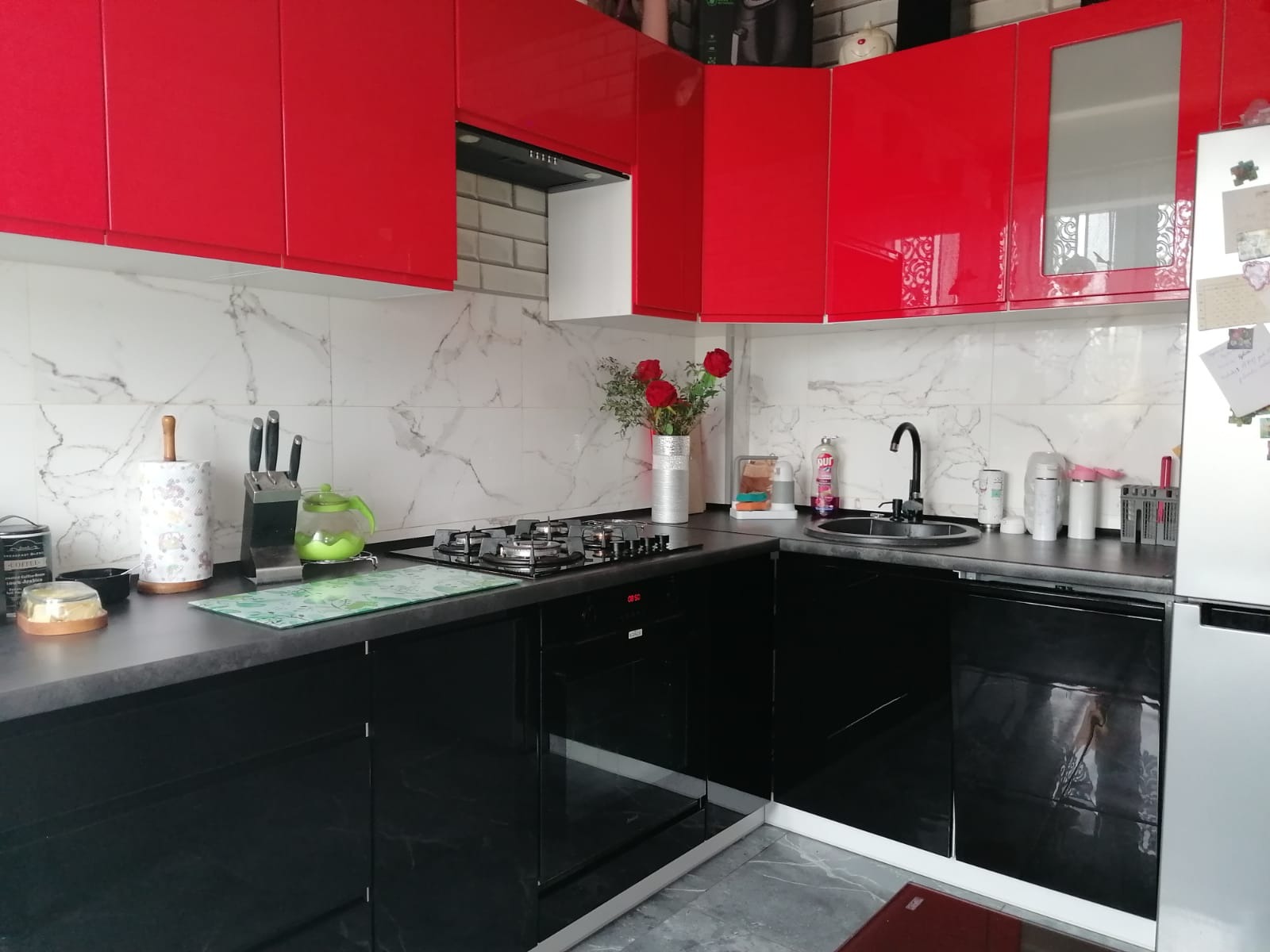 Nowoczesna kuchnia narożna z czerwonymi szafkami górnymi na wysoki połysk i czarnymi szafkami dolnymi, marmurowy backsplash, ozdobiona wazonem z czerwonymi różami.