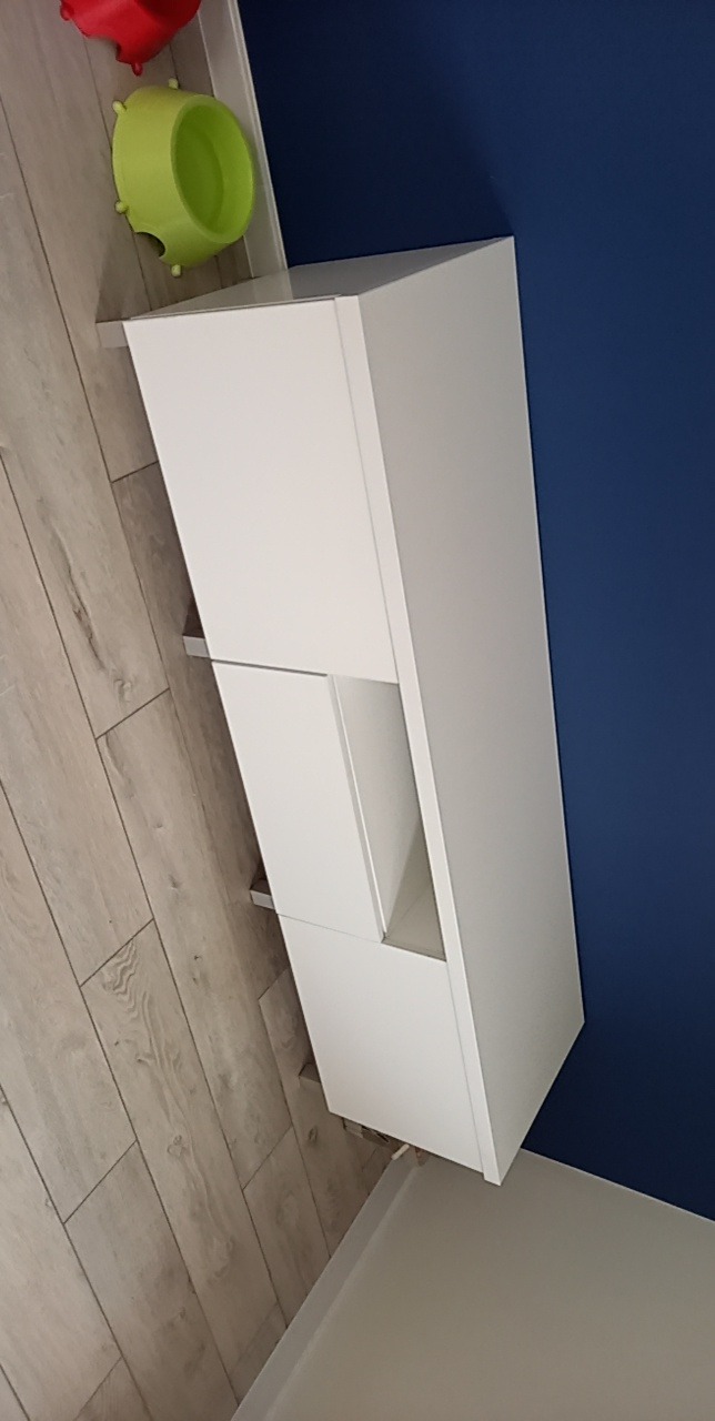 Biała szafka wisząca z asymetrycznymi frontami na tle ciemnoniebieskiej ściany, z widocznymi dziecięcymi zabawkami na górze.