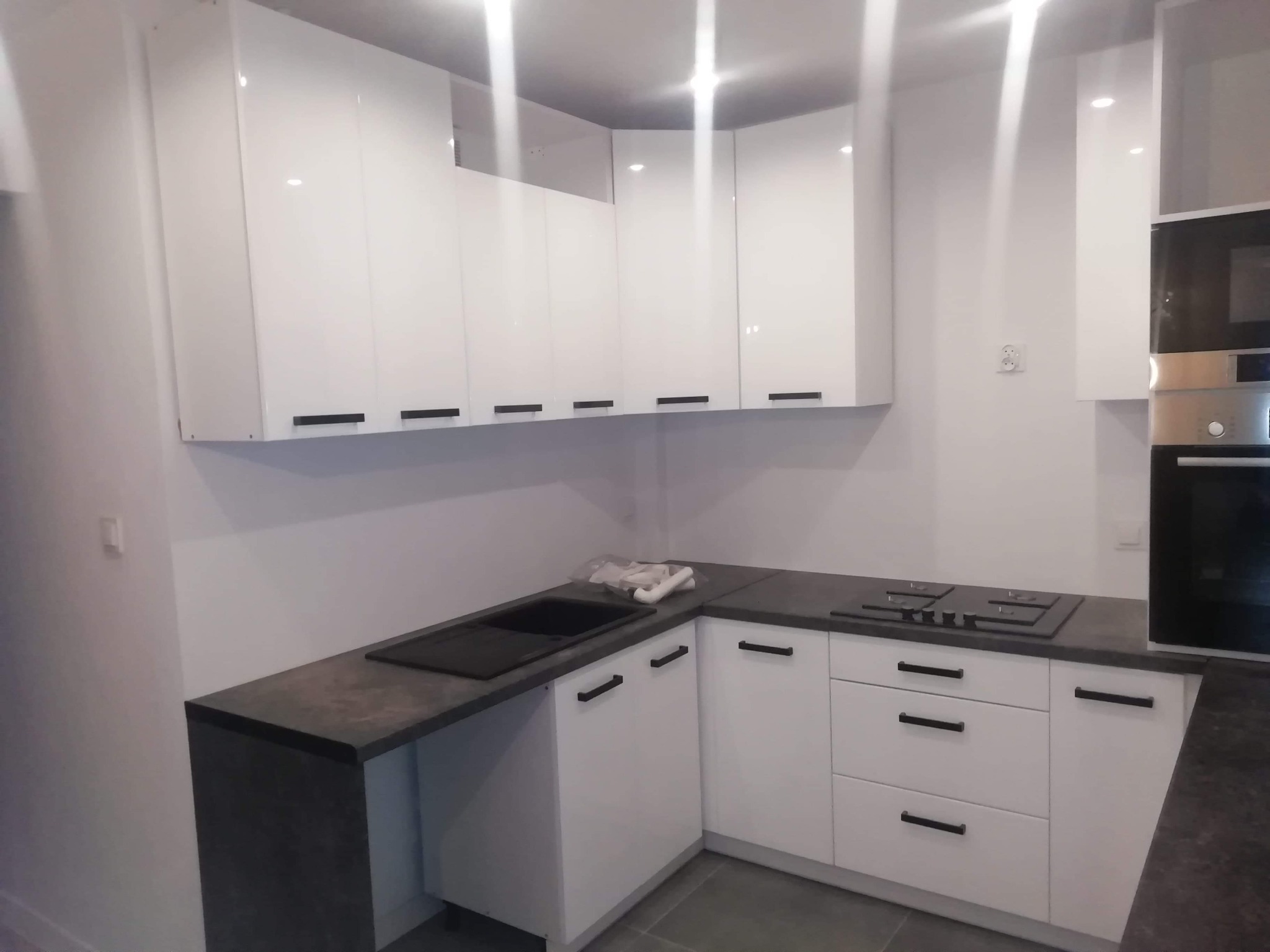 Biała, nowoczesna kuchnia z czarnymi uchwytami, szarym blatem i wbudowaną płytą grzewczą oraz piekarnikiem.