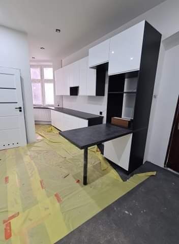 Nowoczesna kuchnia z białymi szafkami górnymi i czarnymi elementami, z rozkładanym blatem na jednej nodze, widoczne zabezpieczenia podłogi folią malarską podczas montażu.