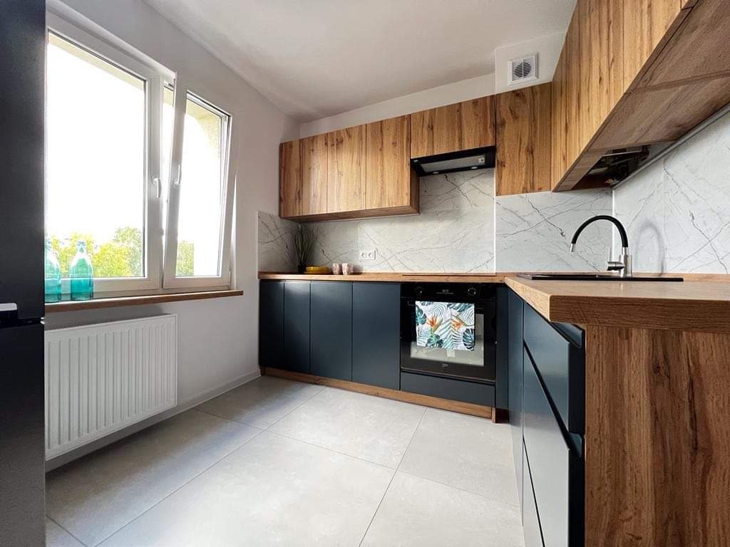 Nowoczesna kuchnia z drewnianymi szafkami górnymi i granatowymi dolnymi, marmurowy backsplash, okno z butelkami na parapecie i ręcznikiem z motywem tropikalnym na piekarniku.