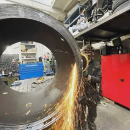 Pracownik w okularach ochronnych szlifuje spaw dużej, metalowej rury przy użyciu szlifierki kątowej, iskry rozbryzgują się w warsztacie.